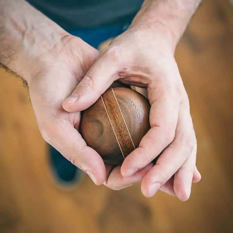 Donkere houten urn met lichtere streep, vastgehouden door twee handen boven houten vloer.