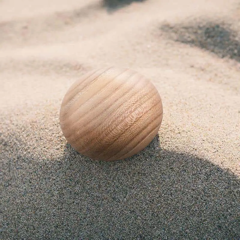 Lichte houten urn met subtiele nerfpatronen, liggend op een zandstrand in zonlicht.