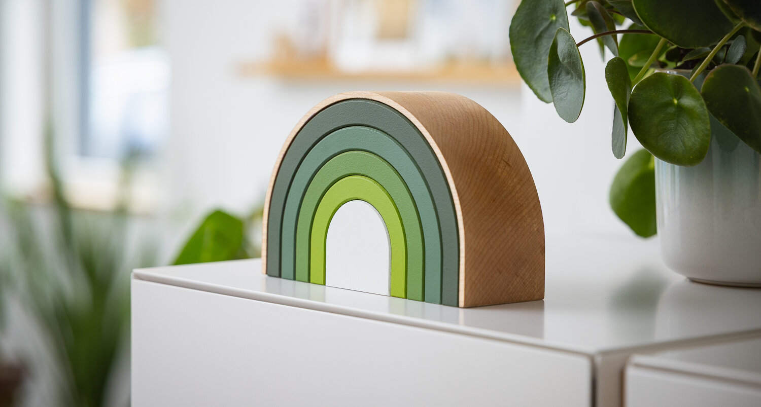 Houten halfcirkelvormige urn Rainbow in groene tinten schuin gefotografeerd op een witte kast, pilea pannenkoekenplant naast de urn