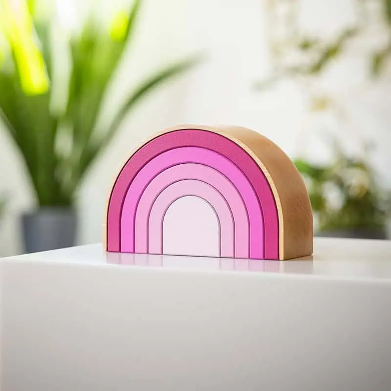 Wooden semicircular Rainbow urn in pink tones, placed on a white surface with plants in the background.