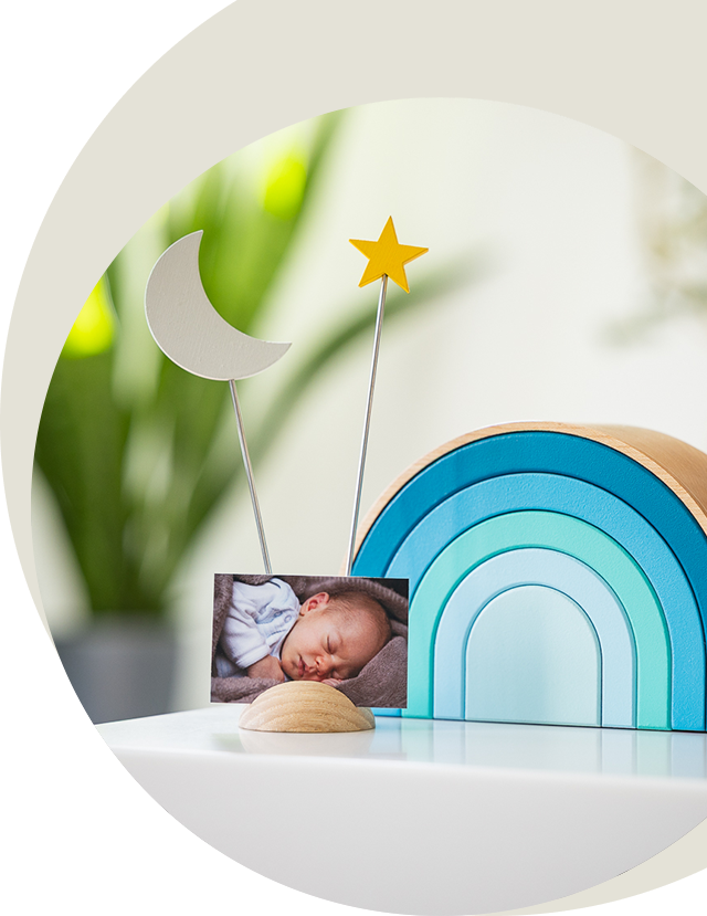Blue rainbow-shaped urn with an integrated photo frame featuring a sleeping baby, adorned with a crescent moon and a golden star, on a white surface.