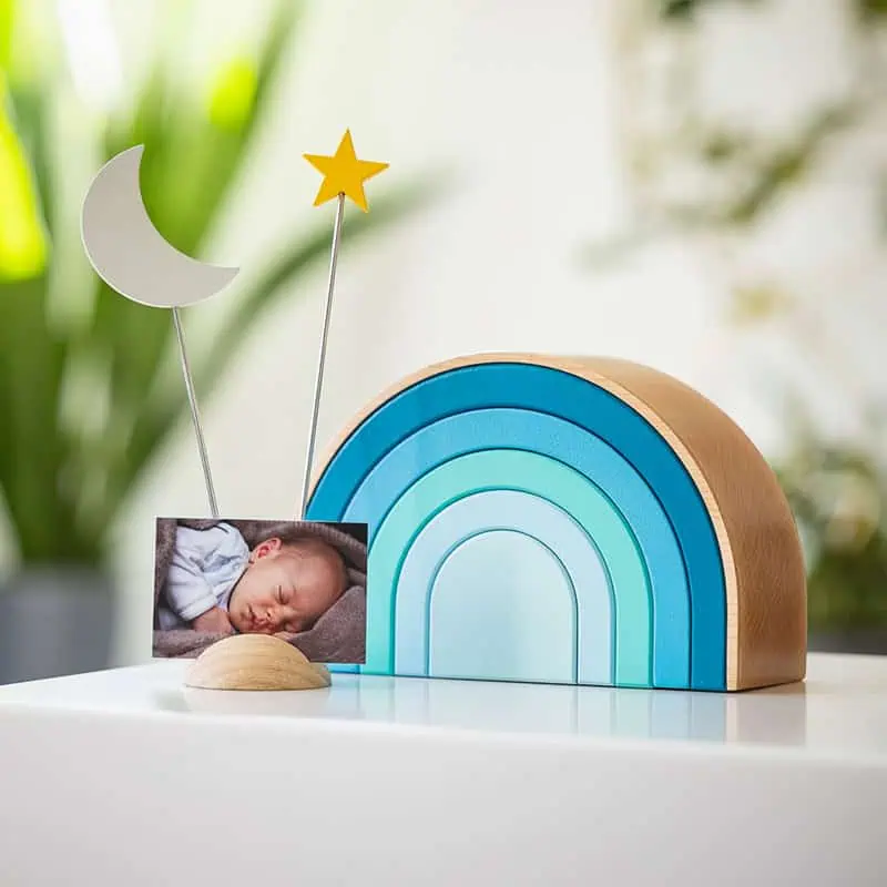 Houten regenboogvormige urn in blauwe tinten met een fotohouder met een foto van een slapende baby, een maansikkel en een gouden ster, op een witte ondergrond met groene planten op de achtergrond
