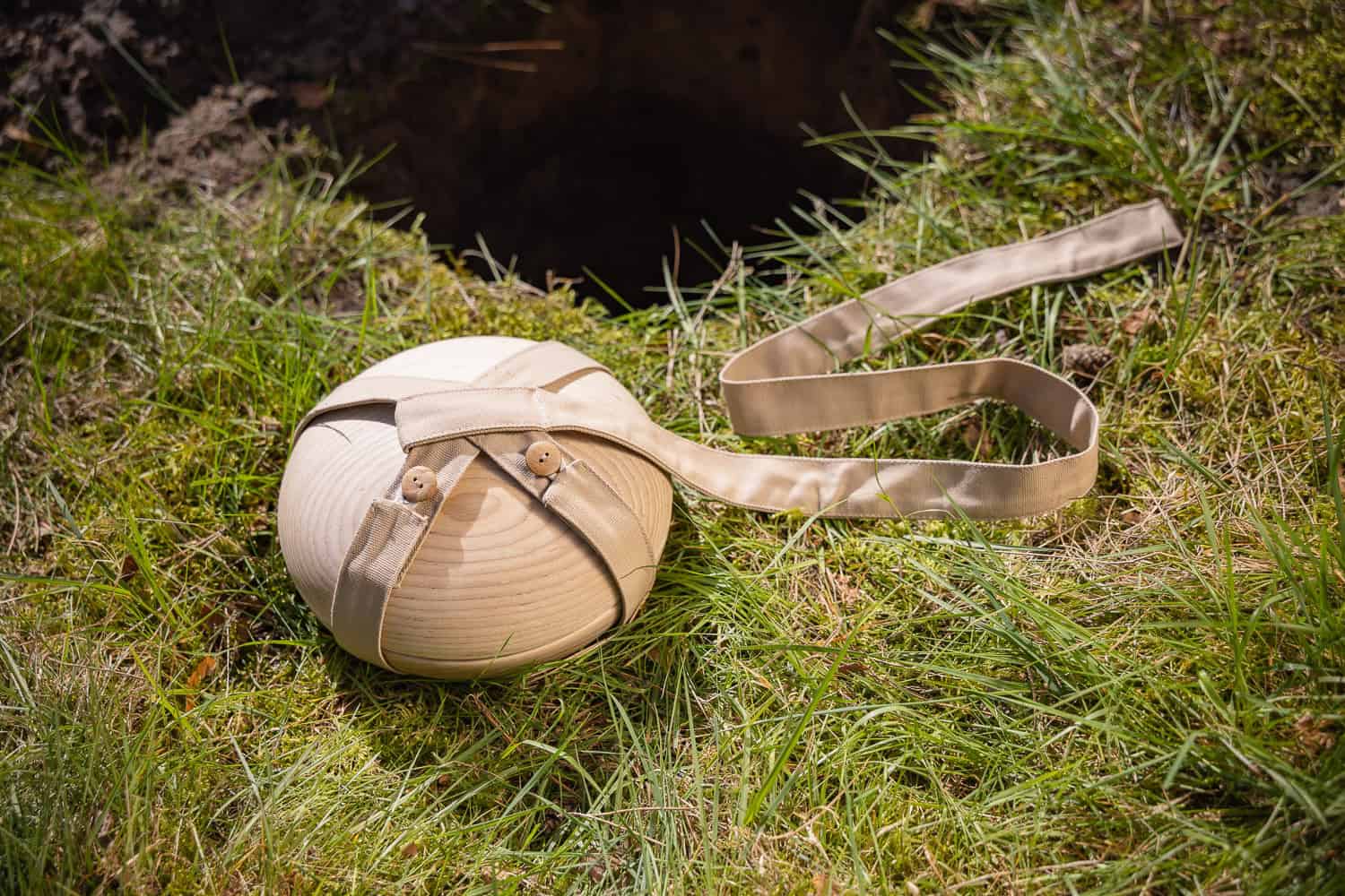 Houten Still urn met stoffen riem en daalband liggend in het gras naast een gegraven kuil