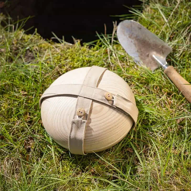 Ronde houten Still urn met beige stoffen riem ligt in groen gras naast een tuinschepje