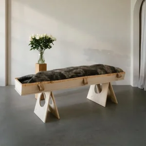 Donkergrijze gevilte wollen wade over een lichaam op een houten baar, met een vaas witte rozen ernaast in een lichte ruimte