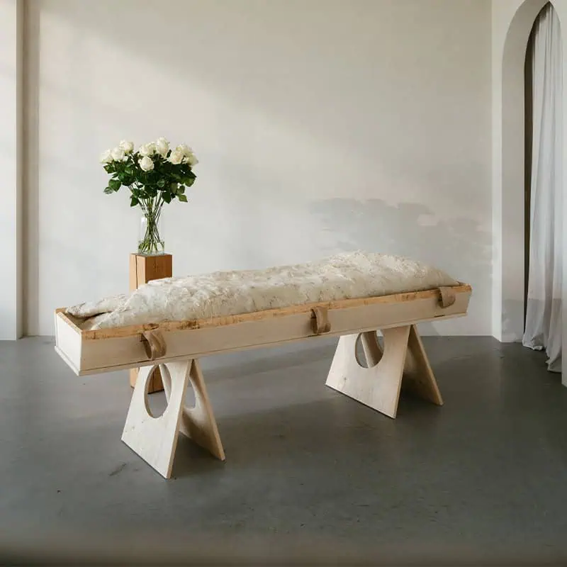 Witte gevilte wollen wade over een lichaam op een houten baar, met een vaas witte rozen ernaast in een lichte ruimte