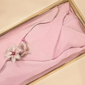 RougeRoze lijkwade van fijn geplooid Egyptisch katoen, gedrapeerd in een lichte houten kist, met een roze magnoliabloem op een takje erop.