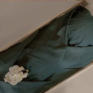 Donkergroen lijkwade van linnen, losjes gedrapeerd in een lichte houten kist, met een witte orchidee erop.