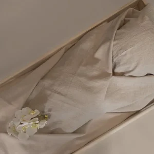 Zand lijkwade van linnen, losjes gedrapeerd in een lichte houten kist, met een witte orchidee erop.