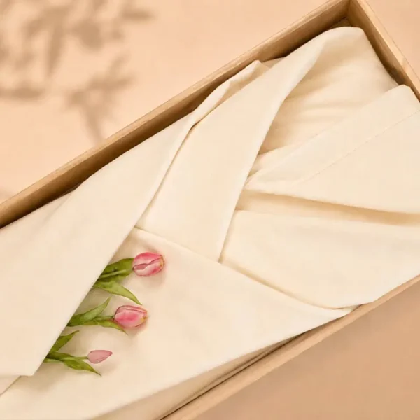 Off-white lijkwade van zachte molton, strak gevouwen in een lichte houten kist, met roze tulpen en groen blad erop.