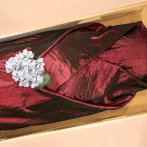 BordeauxRood lijkwade van glanzende taft, gedrapeerd in een lichte houten kist, met een blauwpaarse hortensia erop.