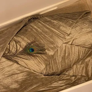 Goud lijkwade van glanzende taft met subtiel cirkelmotief, gedrapeerd in een lichte houten kist, met een pauwenveer erop.