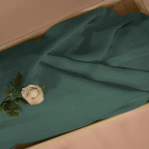Petrolgroene lijkwade van wafelkatoen met wafelstructuur, gedrapeerd in een lichte houten kist, met een cremekleurige roos en groen blad erop.
