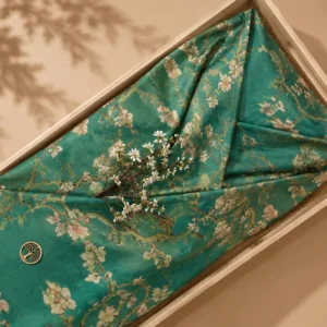 Dubbele wade met een amandelbloesemprint in wit en roze op een zeegroene ondergrond, gevouwen in een lichte houten bak, met een echte bloesemtak en een houten levensboomhanger erop.