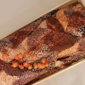 Dubbele wade met een dierenprint van tijger-, luipaard- en slangenmotieven in oranje en bruin, gevouwen in een lichte houten bak, met oranje lampionbloemen en een houten levensboomhanger erop.