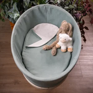 Bovenaanzicht van het Heaven uitvaartmandje in zachtgroen met opengeslagen dekentje en twee knuffels, tussen groene planten