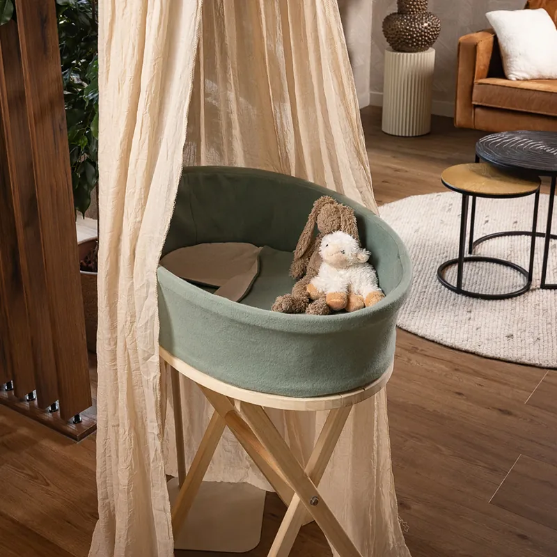 Heaven uitvaartmandje in zachtgroen op een houten standaard achter een beige klamboe, met twee knuffels in de bak