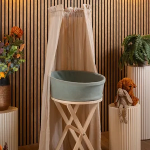 Heaven uitvaartmandje in zachtgroen op een houten standaard onder een klamboe, tegen een houten lattenwand met bloemen en varens ernaast