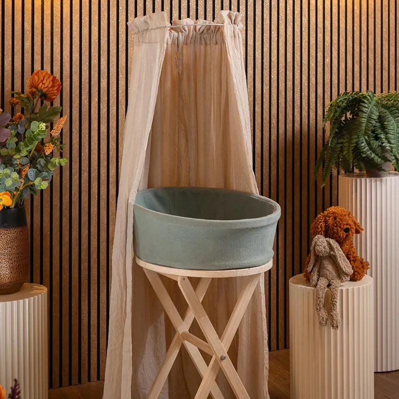 Heaven uitvaartmandje in zachtgroen op een houten standaard onder een klamboe, tegen een houten lattenwand met bloemen en varens ernaast