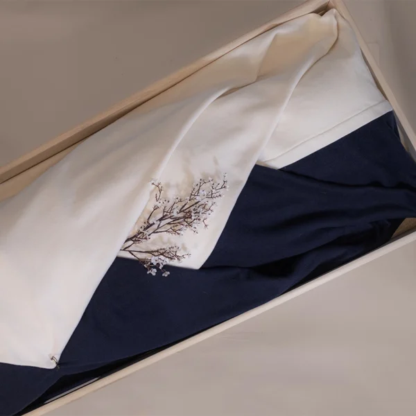 Tweekleurige lijkwade Day and Night in gebroken wit en donkerblauw, gevouwen in een lichte houten bak, met een geweven bloesemtak op het lichte deel.