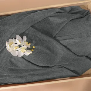 Grijs lijkwade van gebreide wafelstof met duidelijke wafelstructuur, gedrapeerd in een lichte houten bak, met een witte orchidee erop.