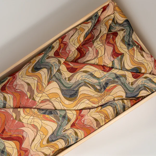 Lijkwade van jacquard met een golvend abstract motief in rood, oker, blauw en groen, gevouwen in een lichte houten bak.