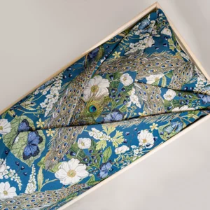 Lijkwade van jacquard met een geweven pauwenprint met witte bloemen en vlinders op een blauwe ondergrond, gevouwen in een lichte houten bak.