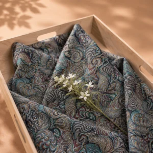 Lijkwade van jacquard met een fijn gestippeld paisleymotief in donkerblauw, turquoise en okerbruin, gevouwen in een lichte houten bak, met een witte bloemtak erop.