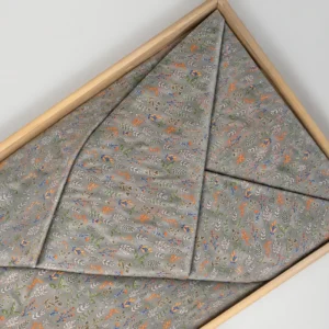 Grijsgroene lijkwade met een fijne botanische print van kleine bloemen en blaadjes in oranje, blauw en groen, gevouwen in een lichte houten bak.