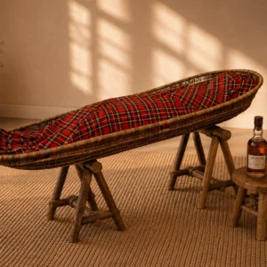 Sfeerfoto van de lijkwade met rode Schotse ruit in een rieten baar op houten schragen, met kaarsen, een ingelijste foto en een fles whisky met glas op een krukje.