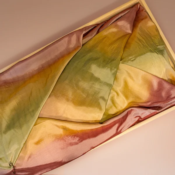 Glanzende lijkwade van taft changeant met een kleurverloop van roze naar olijfgroen en okergeel, gevouwen in een lichte houten bak.