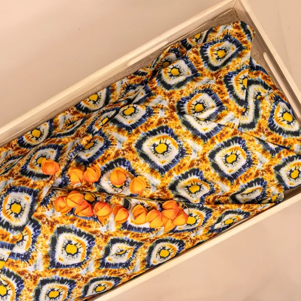 Lijkwade met tie-dye print in oker, geel en blauw ruitmotief, gevouwen in een lichte houten bak, met oranje lampionbloemen erop.