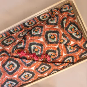 Lijkwade met tie-dye print in oranje en donkergrijs medaillonmotief, gevouwen in een lichte houten bak, met roze bougainvillebloemen erop.